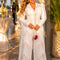A woman models a white long-sleeve jumpsuit with intricate gold embroidery and lace details, styled with gold accessories and a small red handbag in an elegant outdoor setting.