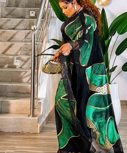 Woman in a green and black traditional outfit standing in a home setting with plants and stairs.